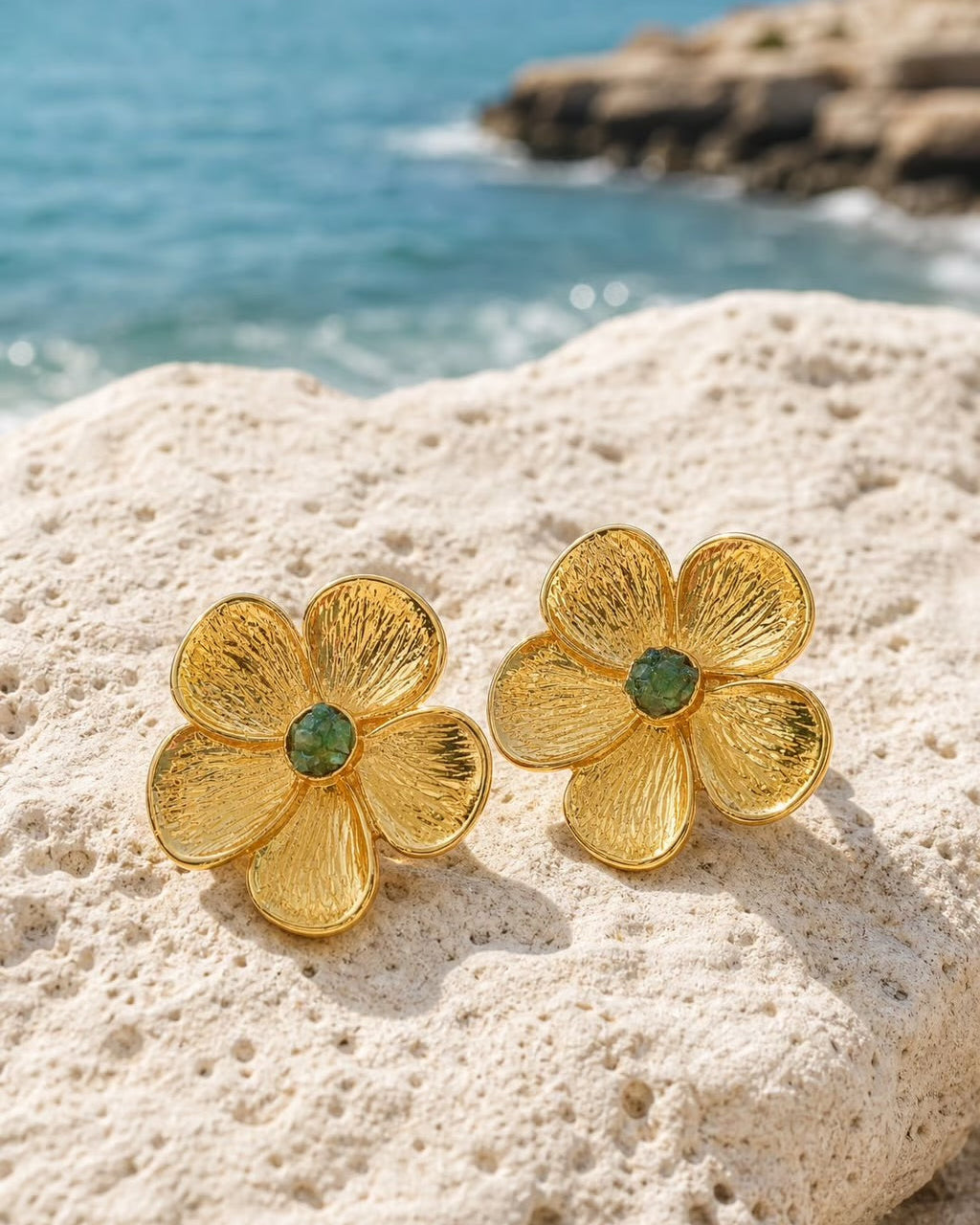 A pair of gold flower-shaped stud earrings with small green gemstone centers, resting on a textured white rock with a blurred ocean background.