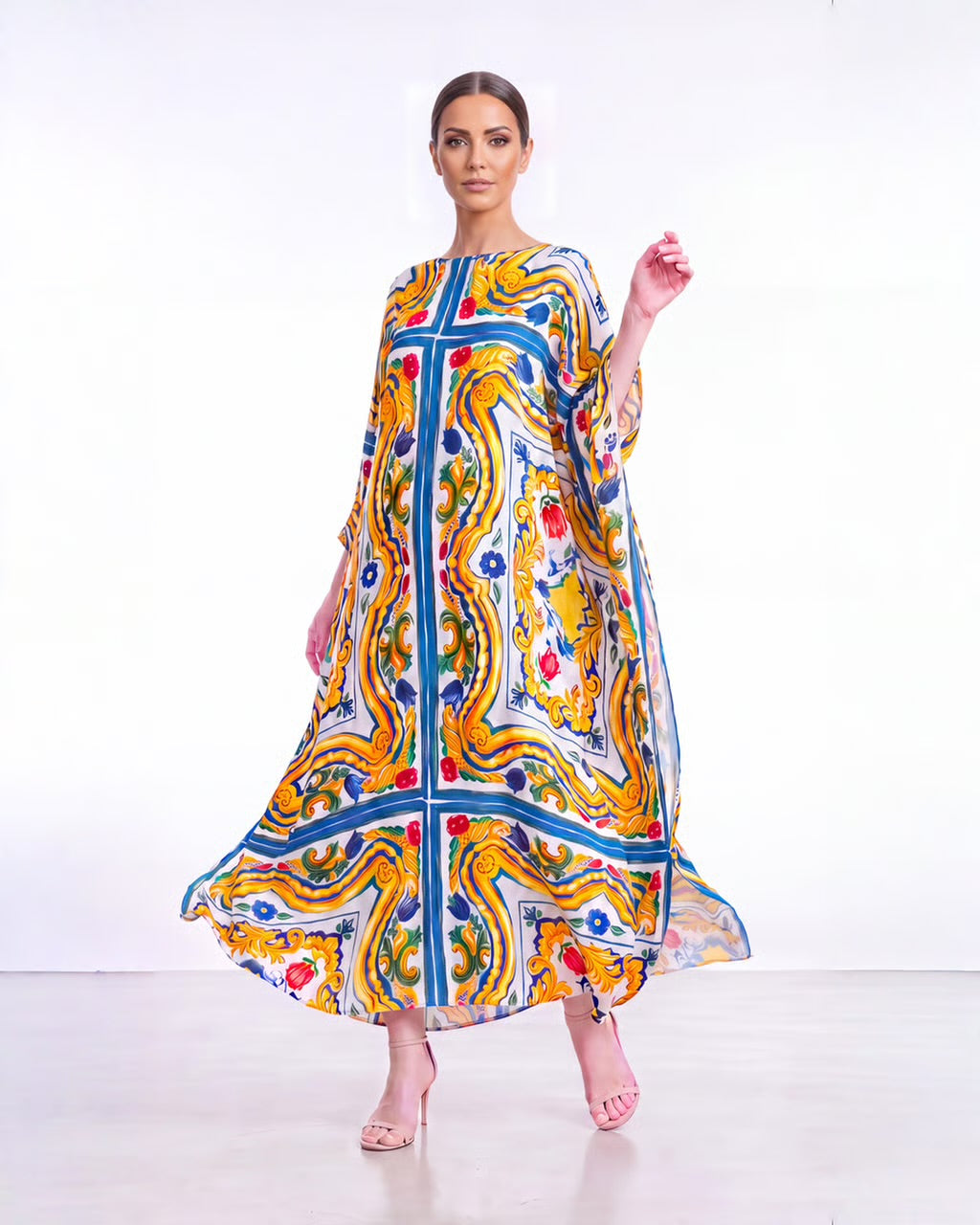 A woman modeling a long, flowing kaftan dress with a vibrant, multicolored abstract print featuring yellow, blue, orange, and red swirls on a white base, set against a plain white studio background.