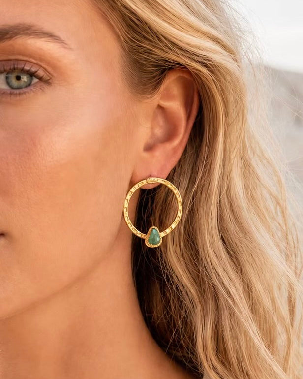 A close-up of a woman wearing a textured gold hoop earring with a small green teardrop gemstone dangling from the side.