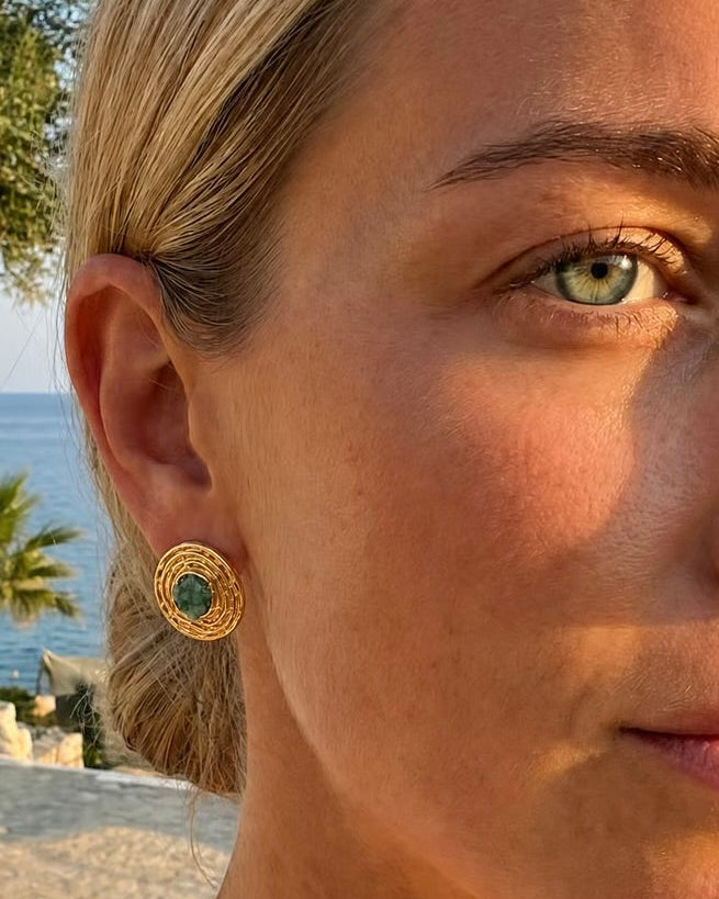 A close-up of a woman's ear wearing a gold circular stud earring with a green circular center, set against a blurred outdoor background of water and trees.