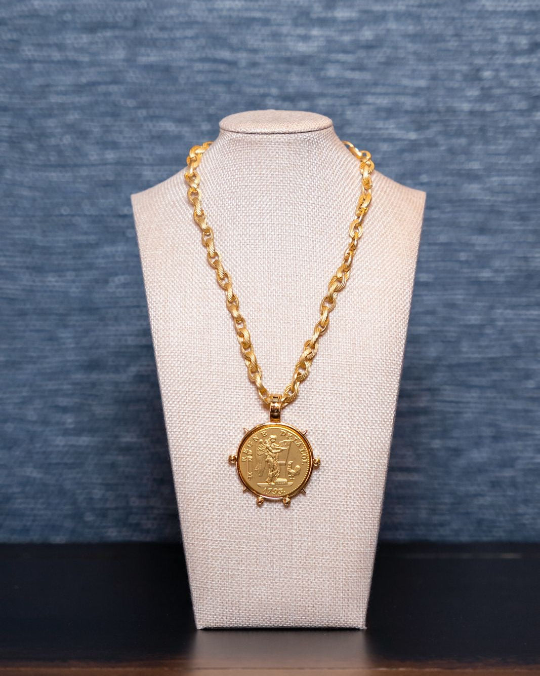 Gold necklace with a pendant on a mannequin bust against a blue textured background