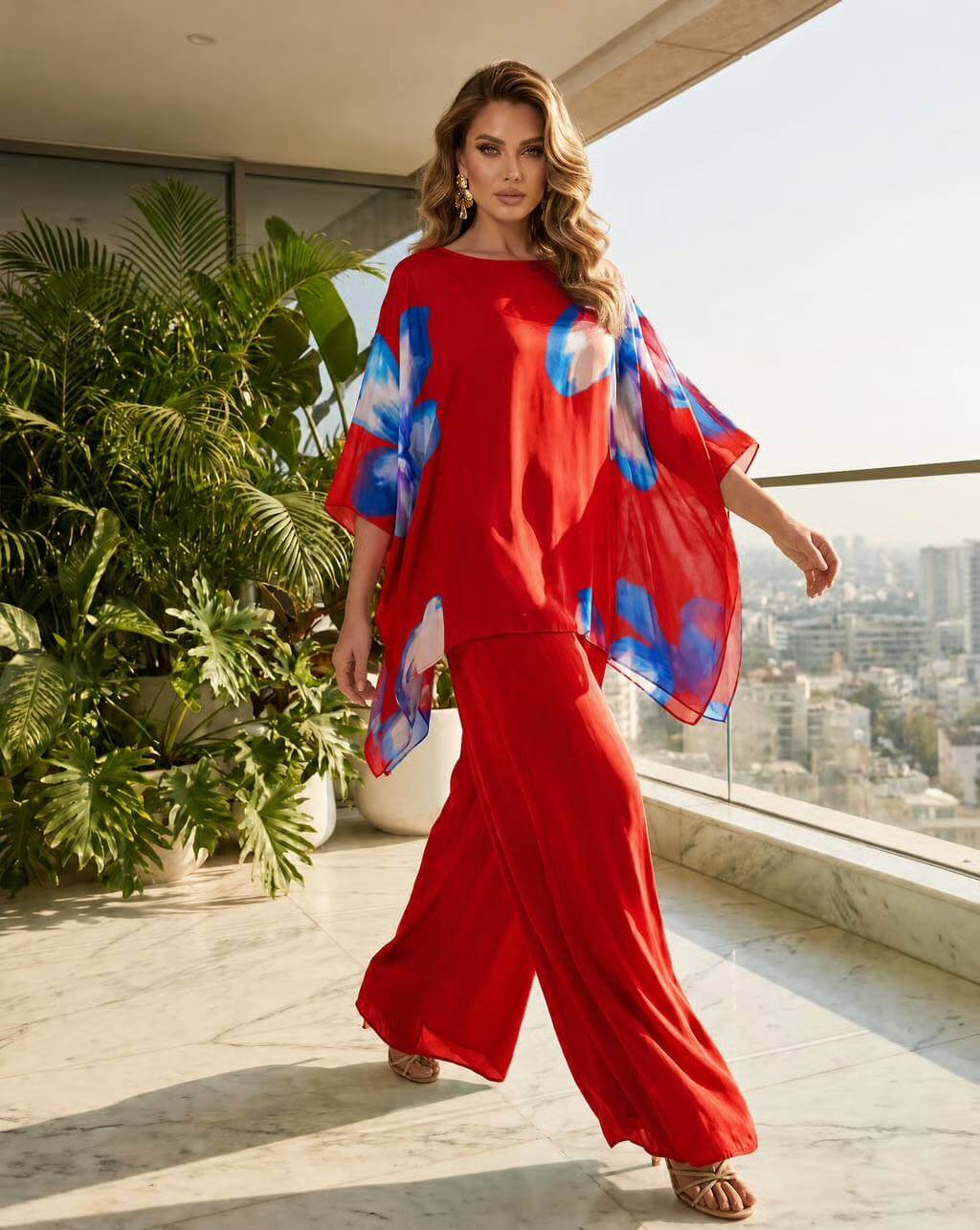 A woman models a vibrant red two-piece lounge set consisting of a short-sleeve top with blue and white tie-dye patterned sleeves and matching wide-leg pants, set against a modern balcony backdrop with plants and a city view.