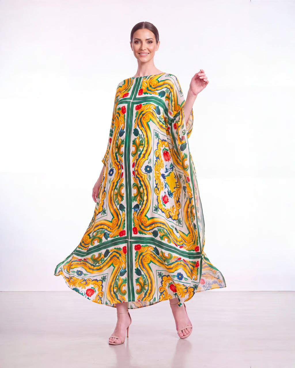 A woman modeling a long, flowing kaftan dress with a vibrant yellow, green, and white paisley pattern, standing against a plain white studio background.