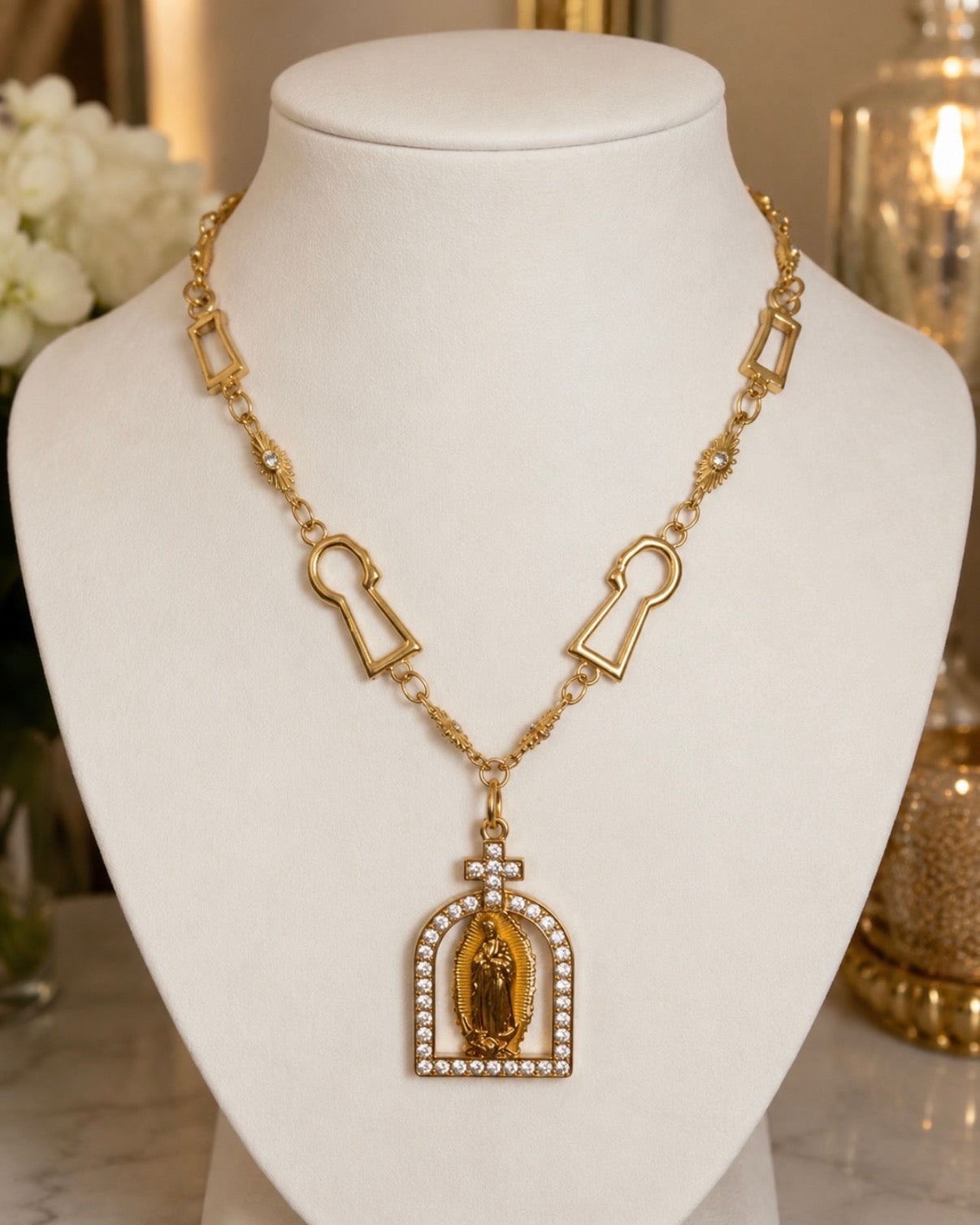 Gold necklace with keyhole and arched link details, featuring a central pendant with a religious figure surrounded by small white stones, displayed on a white mannequin bust.