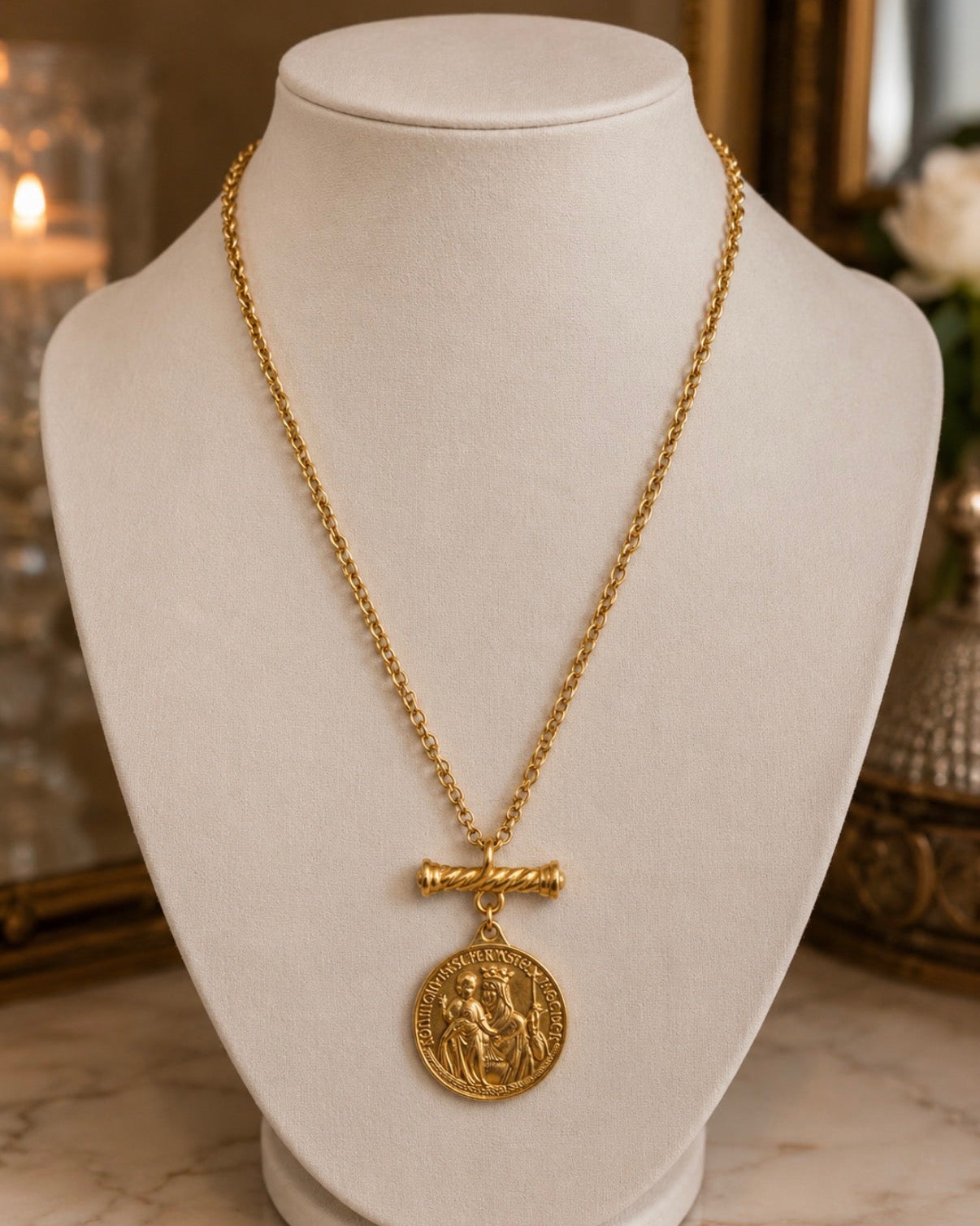 A gold necklace with a round coin pendant and a twisted bar clasp, displayed on a white jewelry bust against a softly blurred indoor background.