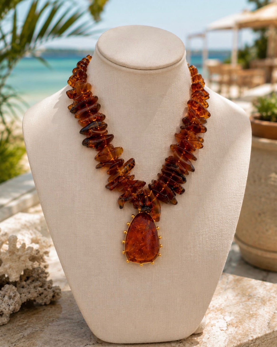 A chunky amber necklace with a large teardrop pendant, displayed on a cream fabric mannequin bust. The background is a blurred tropical beach scene with turquoise water, palm leaves, and outdoor patio furniture.