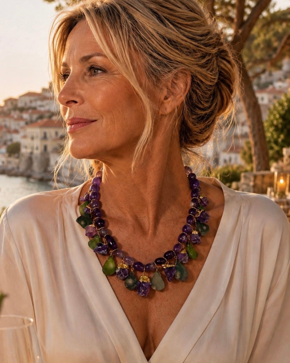 A woman wearing a chunky multi-strand statement necklace made of purple and green beads, posing outdoors near a waterfront at sunset.