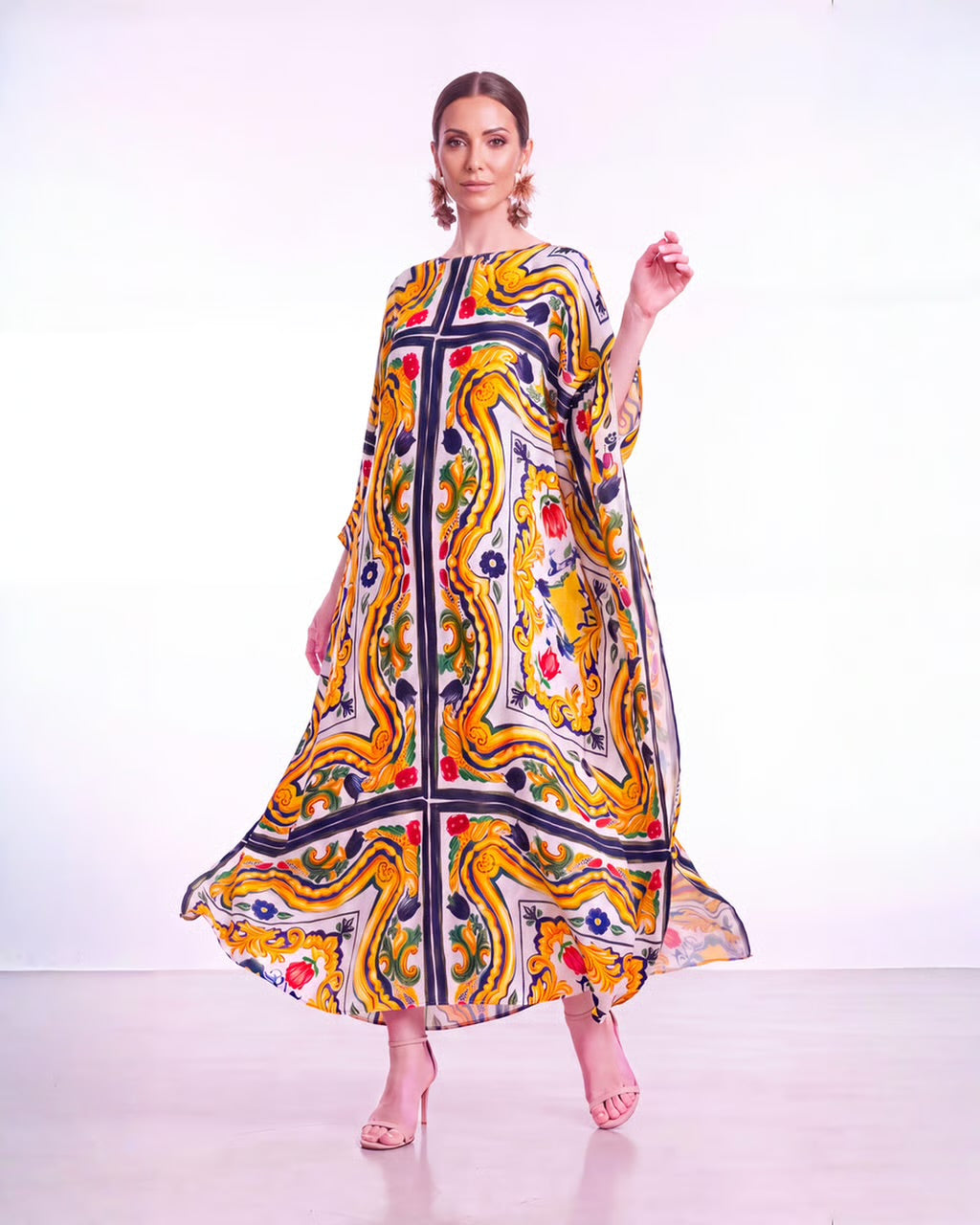 A woman modeling a long, flowing kaftan dress with a vibrant abstract print in yellow, orange, navy, green, and red on a white background, set against a plain white studio backdrop.
