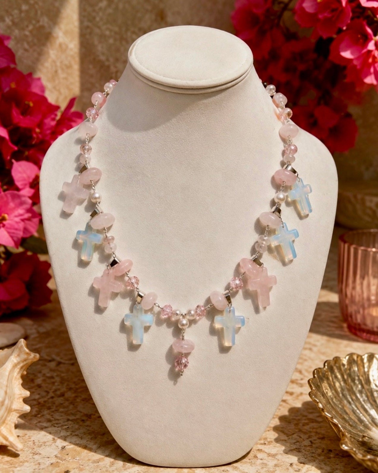 A beaded necklace with light pink and pale blue cross charms displayed on a cream-colored jewelry bust, with pink flowers and decorative props in the background.