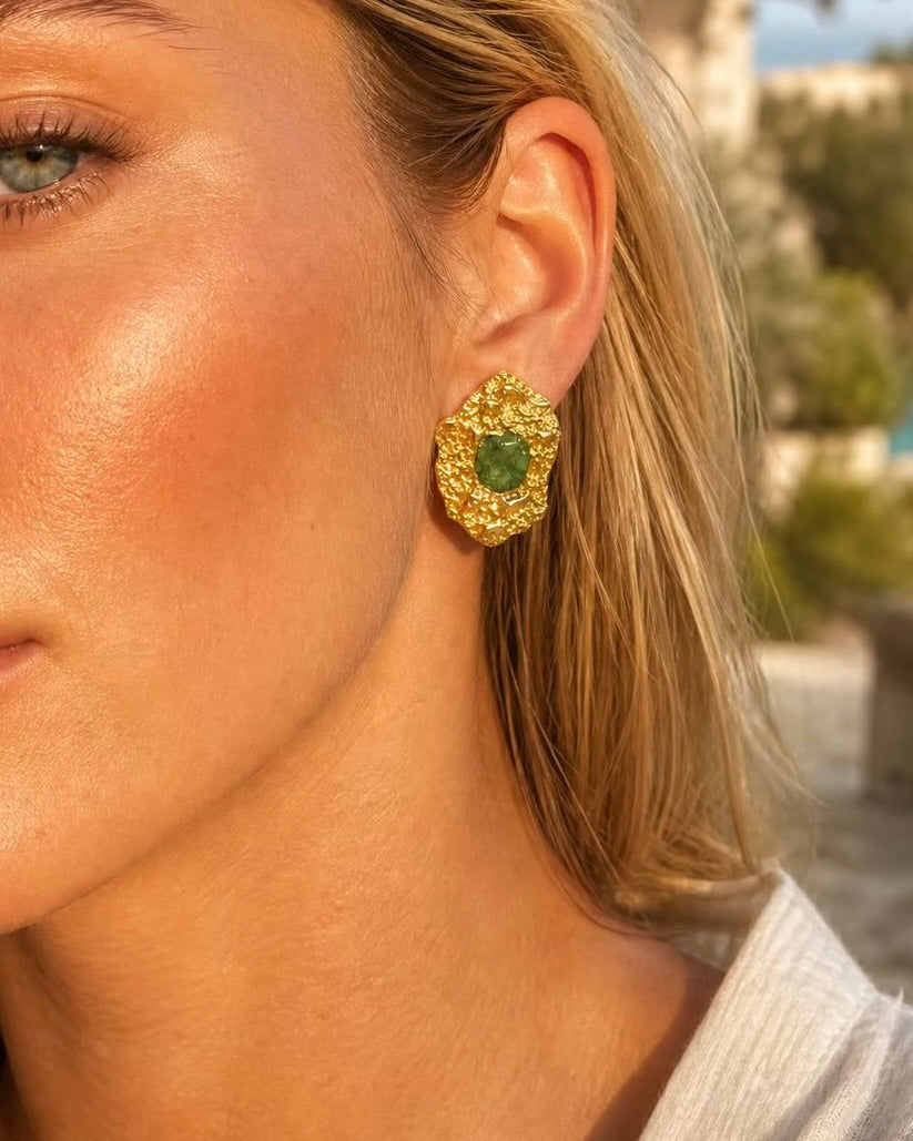 A close-up of a woman wearing a textured gold statement earring with a central green gemstone, set against a softly blurred outdoor background.