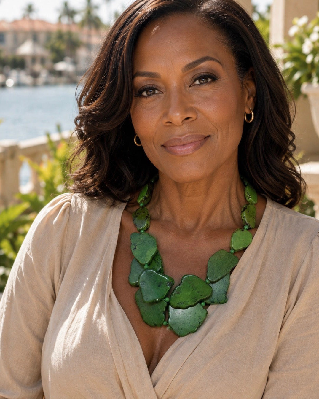 A woman with dark hair wearing a beige blouse and a chunky green stone necklace, photographed outdoors with a waterfront and buildings in the background.