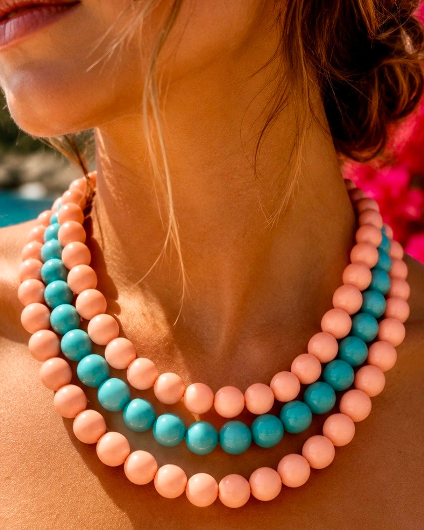 A close-up lifestyle shot of a woman wearing a three-strand beaded necklace with alternating pastel peach-pink and turquoise blue beads.