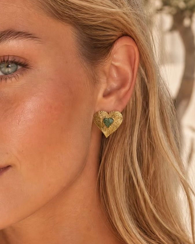 A close-up of a woman's ear wearing a textured gold heart-shaped stud earring with a green heart in the center.