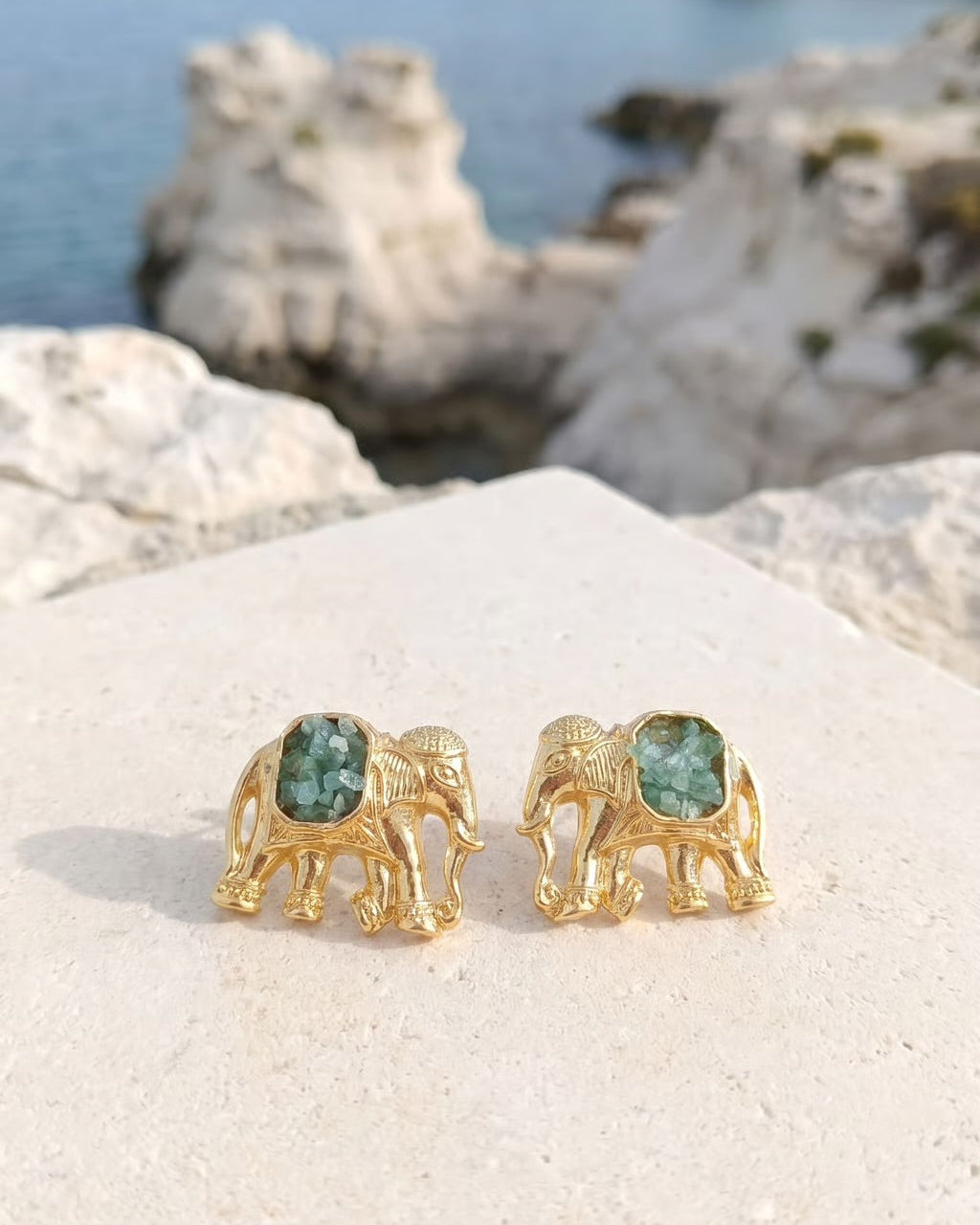 A pair of gold elephant stud earrings with green gemstone backs, displayed on a light stone surface with a blurred coastal background.