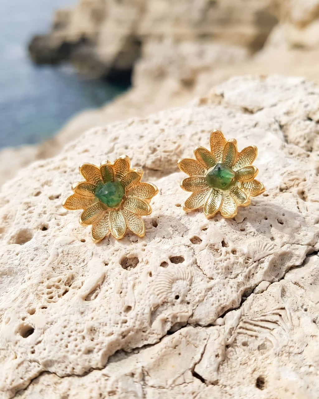 A pair of gold flower-shaped earrings with green gemstone centers, resting on a textured beige rock with a blurred coastal cliff and sea in the background.