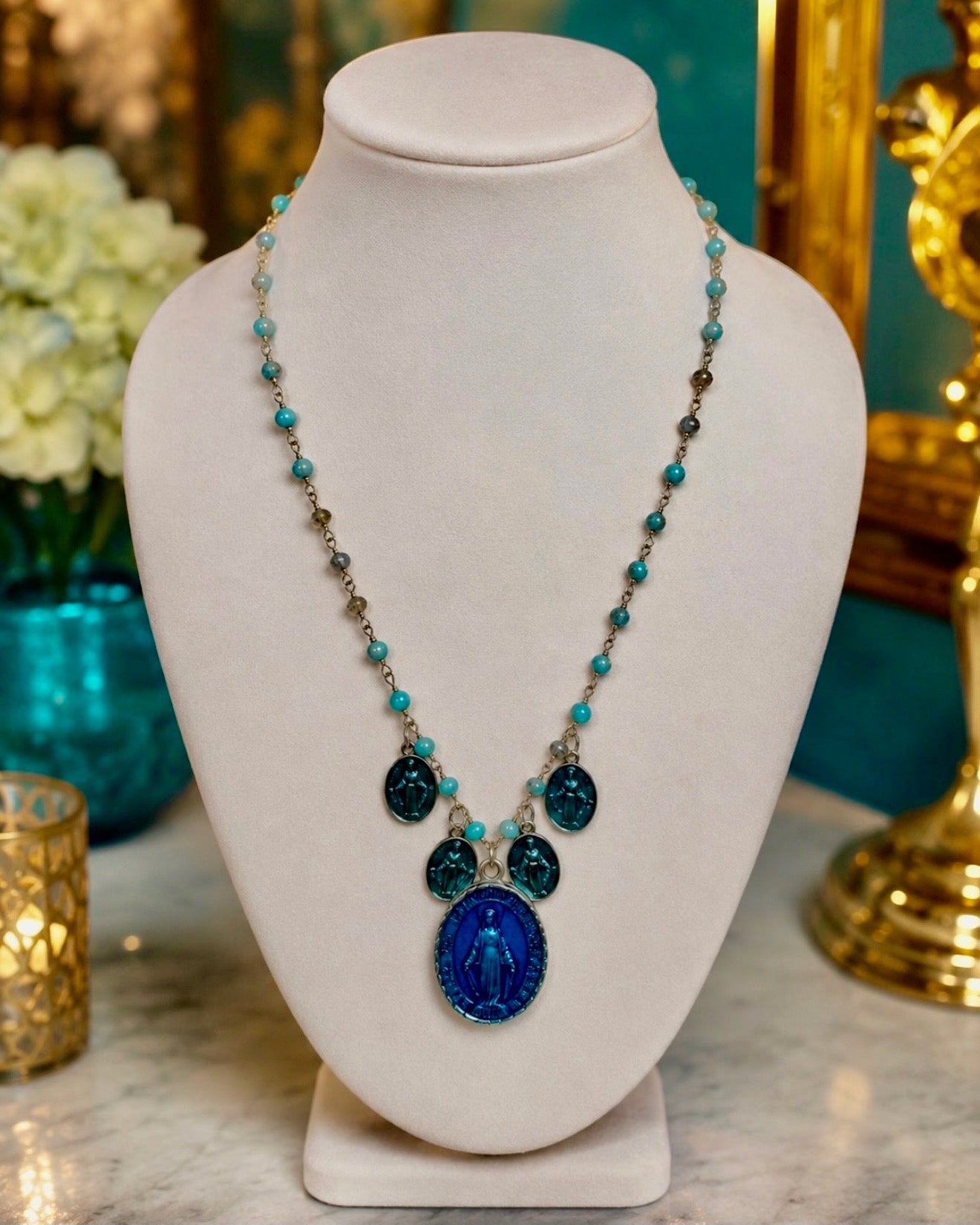 A beaded necklace with a large blue oval pendant featuring a religious figure, displayed on a white jewelry bust. The necklace features turquoise, dark blue, and metallic beads with silver-toned links.
