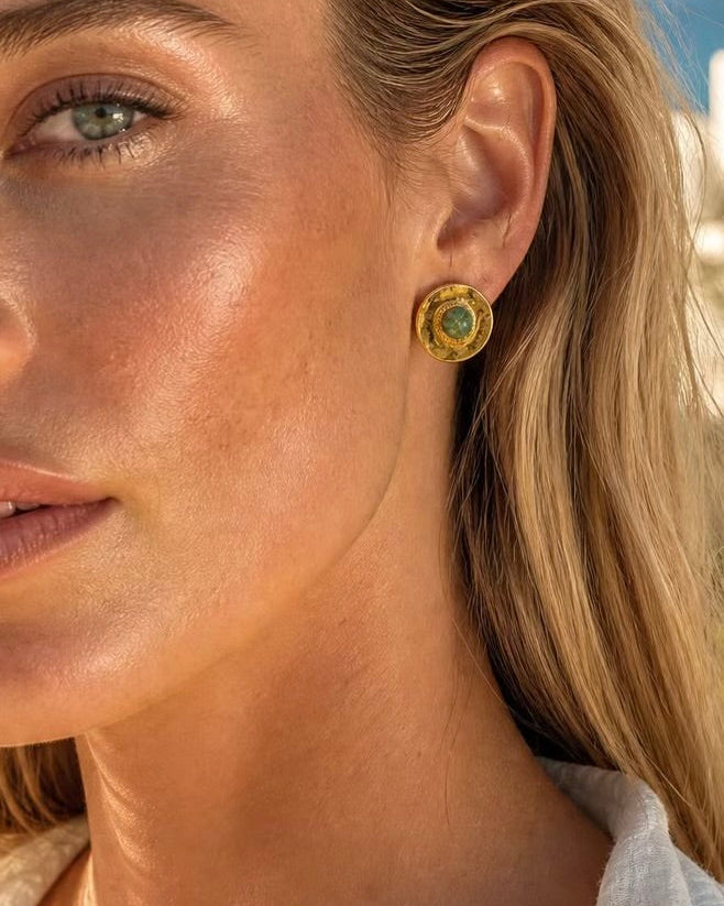 A close-up of a woman's ear wearing a small gold stud earring with a round green stone, set against a softly blurred outdoor background.