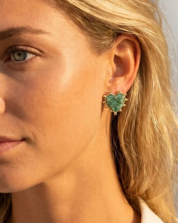 A close-up of a woman's ear wearing a textured green heart-shaped stud earring with a gold-toned setting.