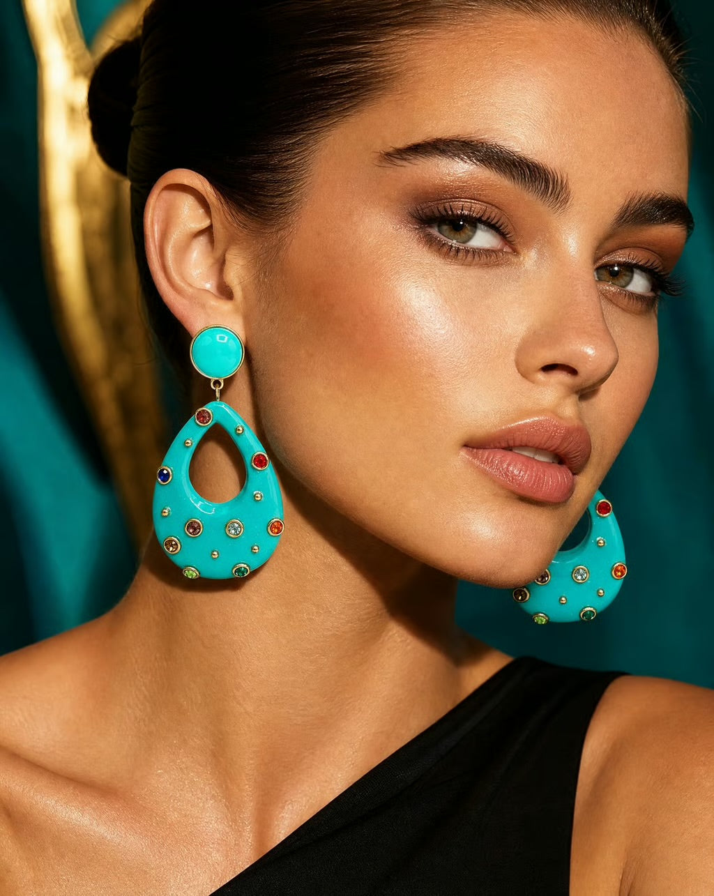 A close-up of a model wearing a pair of large turquoise teardrop-shaped statement earrings adorned with small gold studs and red and green rhinestone accents, set against a dark teal background.