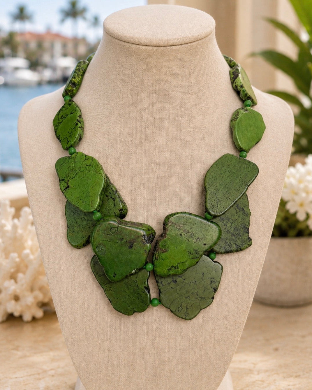 A chunky statement necklace made of irregular green stone pieces, displayed on a beige fabric mannequin bust with a blurred waterfront background.