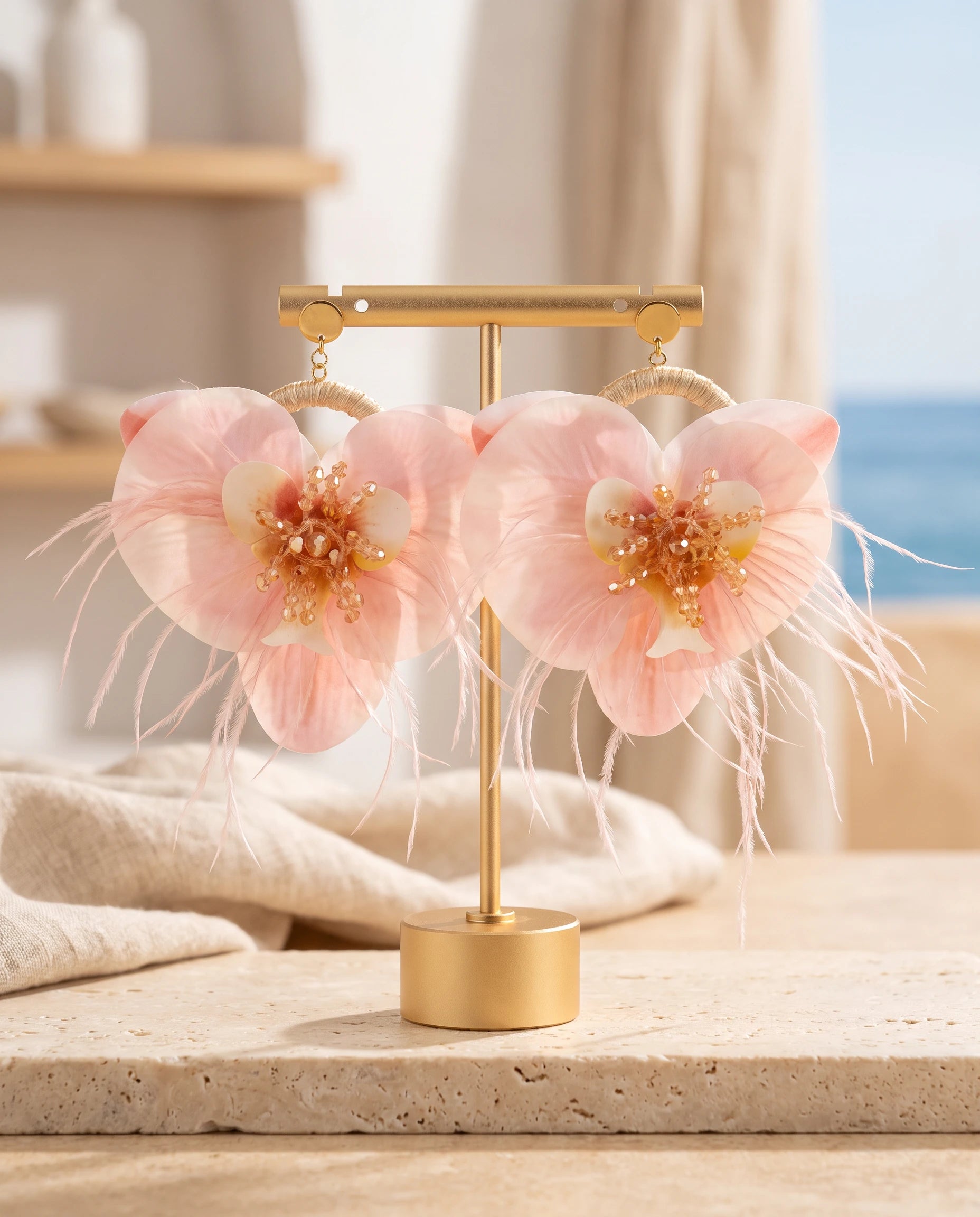A pair of dangling flower earrings displayed on a gold stand. The earrings feature large light pink flower petals with yellow and white centers and wispy feather accents, shown against a softly blurred beachside interior background.