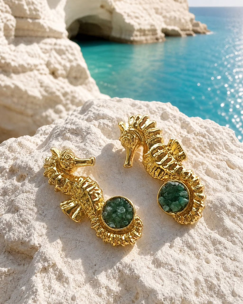 A pair of gold seahorse-shaped earrings with round green gemstone centers, resting on a white rock with a turquoise sea and rocky coastline in the background.