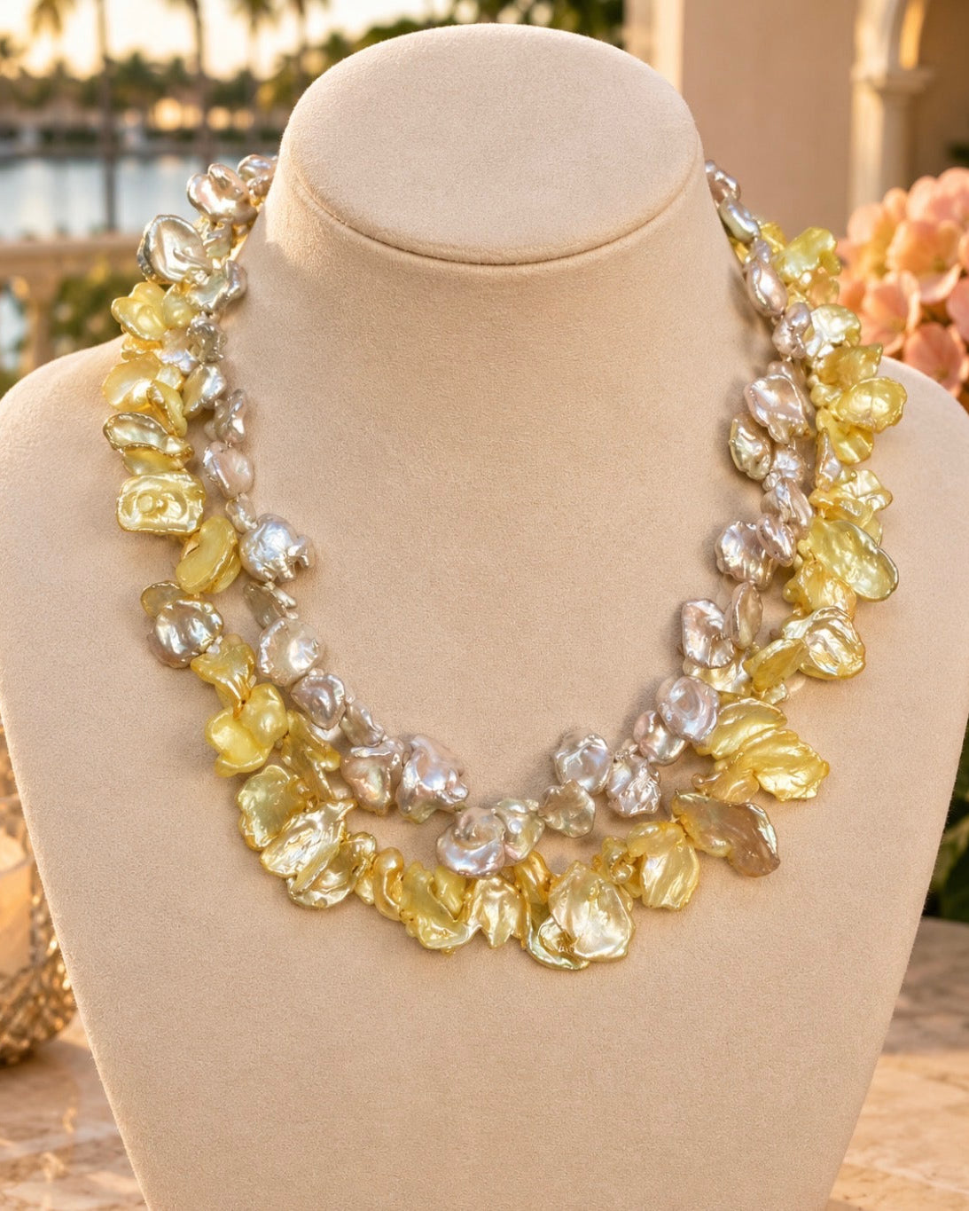 A statement collar necklace made of irregular baroque pearls in yellow, silver, and white tones, displayed on a beige fabric jewelry bust.