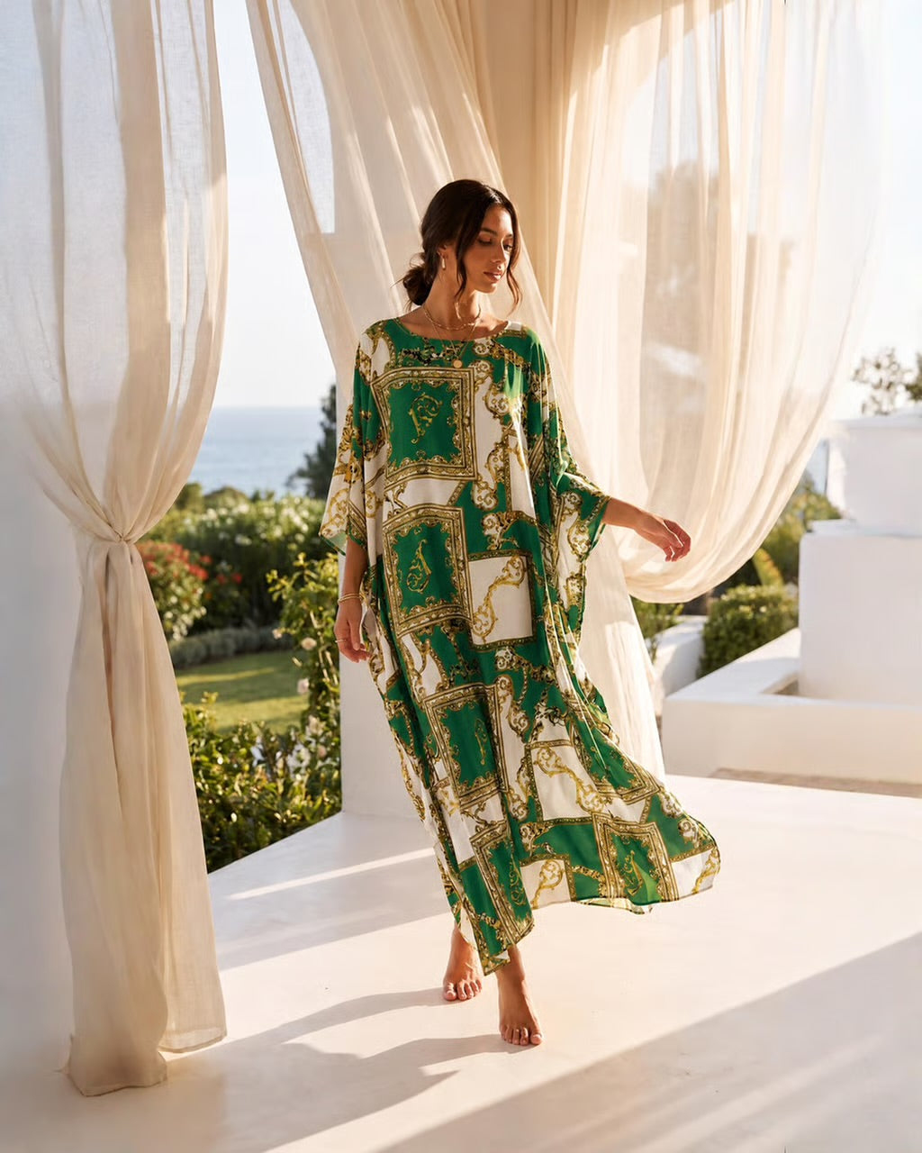 A woman wearing a green and gold baroque-patterned kaftan dress walks barefoot on a sunlit white terrace framed by flowing cream-colored curtains, with a garden and sea in the background.