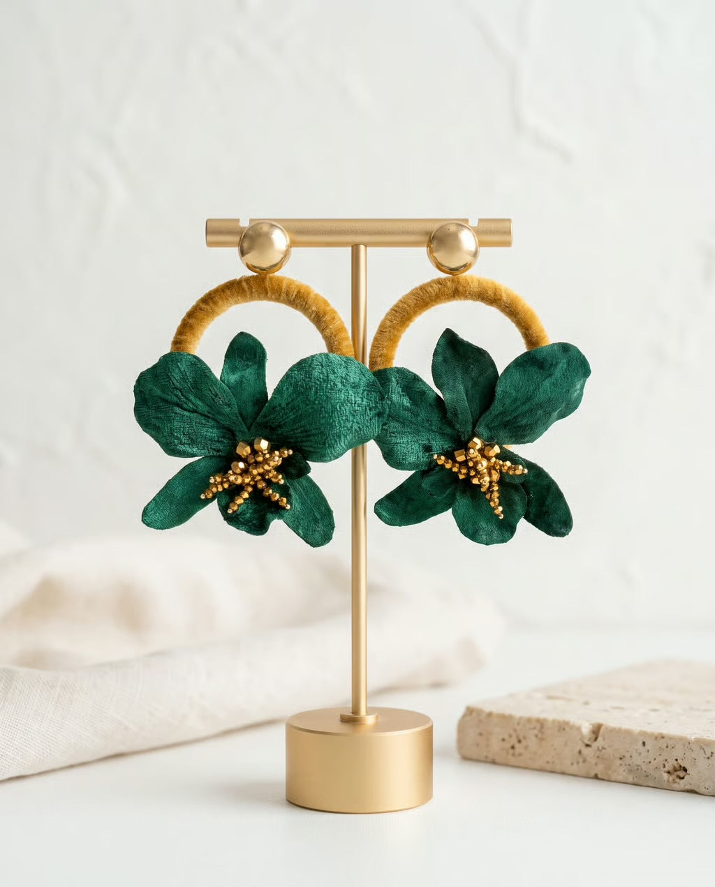 A pair of gold hoop earrings with large green flower charms and gold bead centers, displayed on a gold stand against a soft white background.