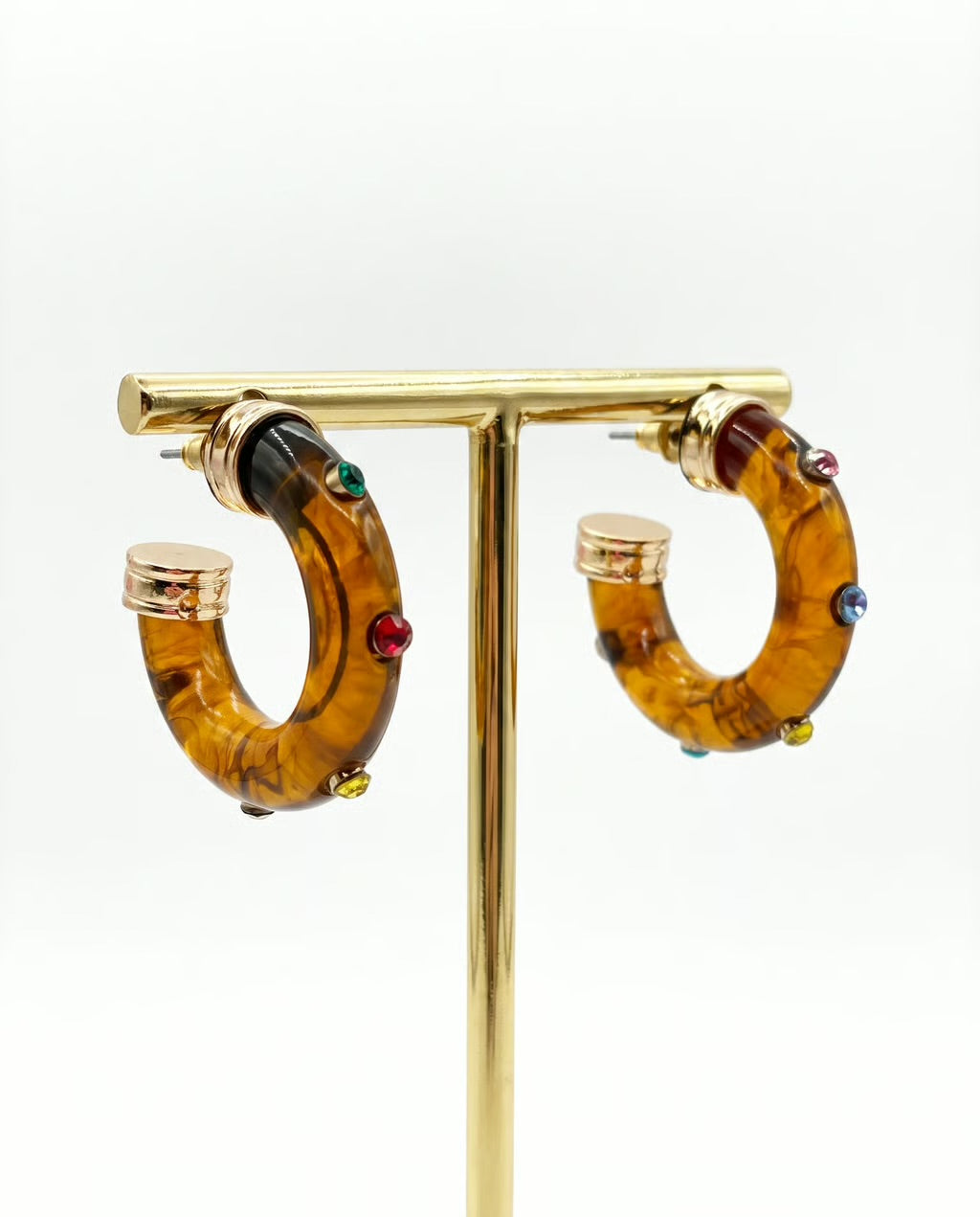 A pair of gold hoop earrings with amber-brown tortoiseshell-like resin sections and small multicolored gemstone accents, displayed on a gold stand against a white background.