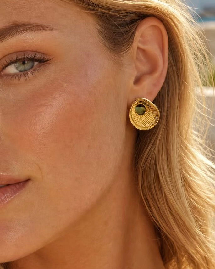 A close-up of a woman's ear wearing a textured gold circular stud earring with a small green stone accent.