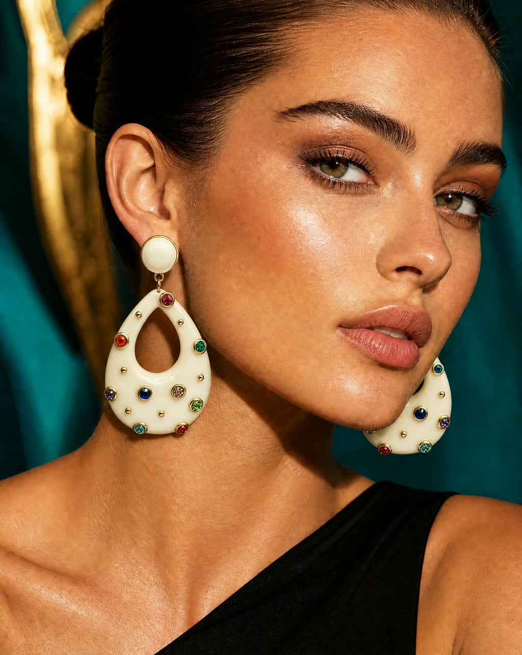 A close-up of a woman wearing large cream-colored teardrop earrings adorned with colorful rhinestones, set against a dark teal background.
