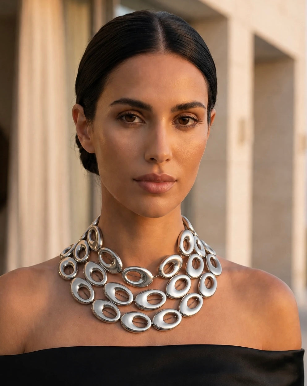 A close-up portrait of a woman wearing a chunky, multi-strand silver statement necklace made of interlocking oval links, paired with a black off-the-shoulder top against a warm-toned architectural background.