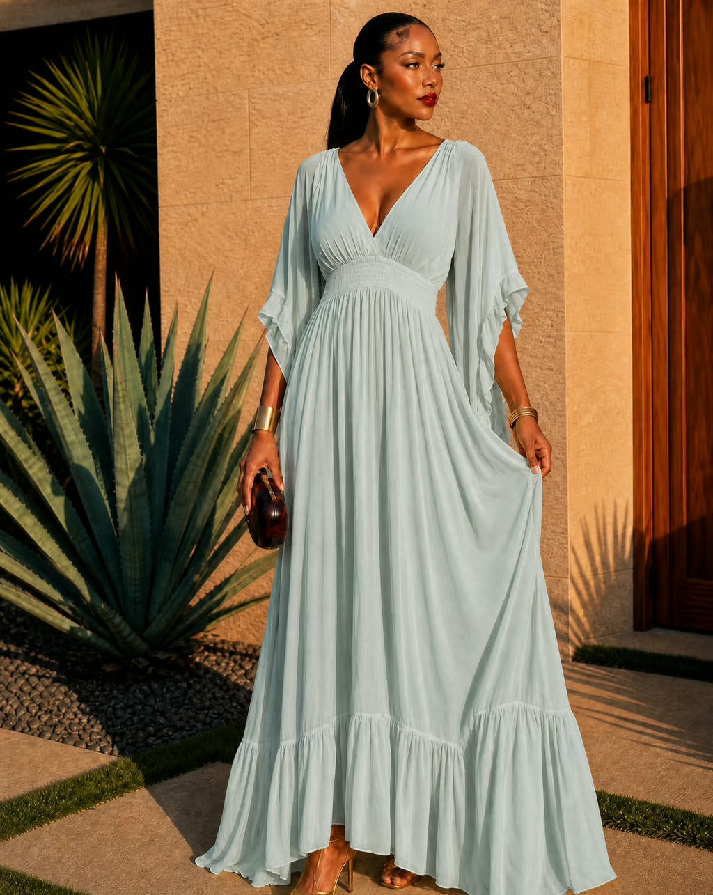 A woman wearing a flowing light blue maxi dress with a deep V-neckline and draped sleeves, standing outdoors in front of a beige stucco wall and agave plant.