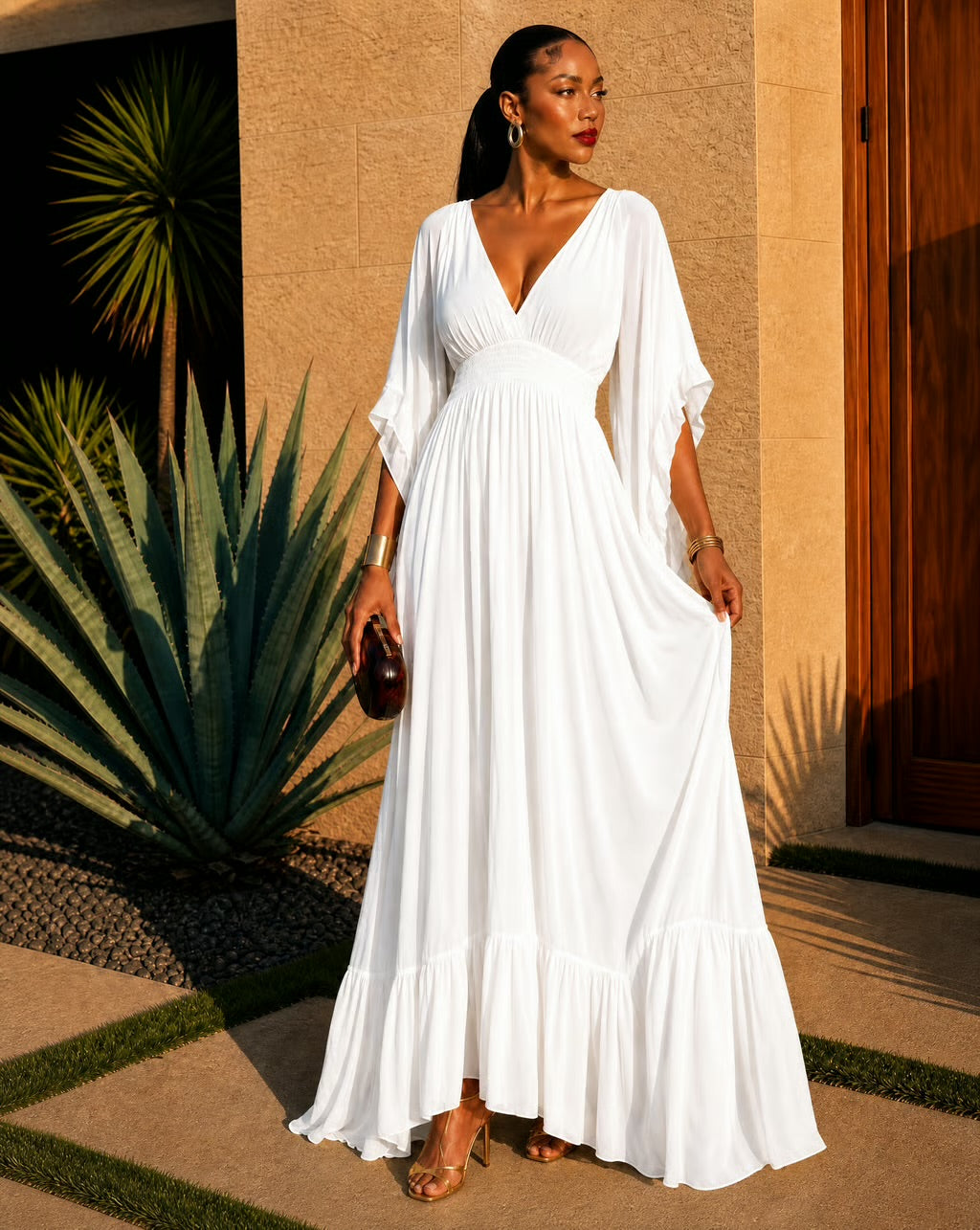 A woman wearing a white maxi dress with a deep V-neckline, elbow-length flutter sleeves, and a tiered ruffled hem, standing outdoors in front of a beige stucco wall and agave plant.