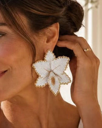 A woman wearing large white flower-shaped statement earrings with gold trim, shown in a bright outdoor setting with a blurred background of water and architecture.