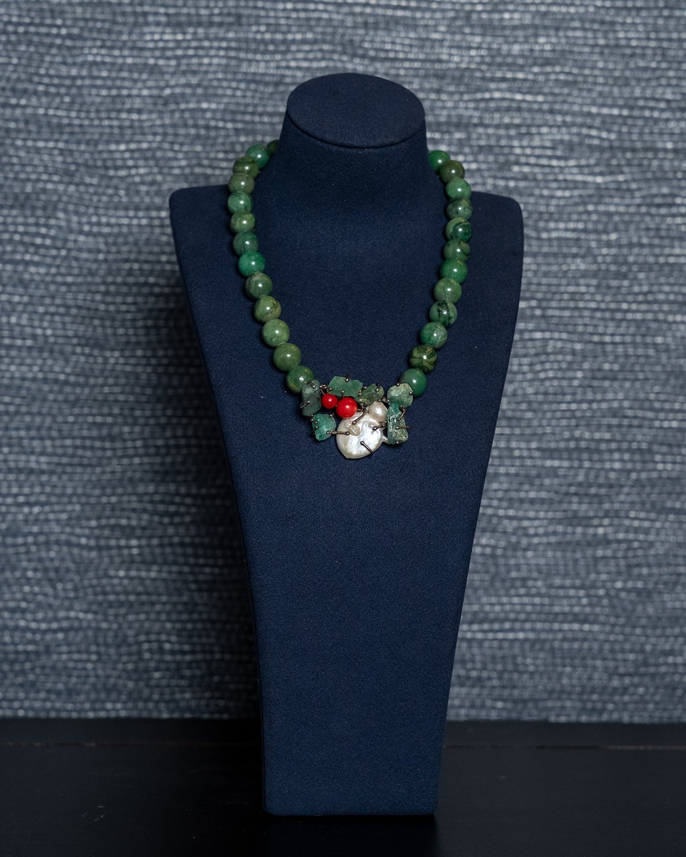 Green beaded necklace with a red and white pendant on a blue stand against a gray textured background