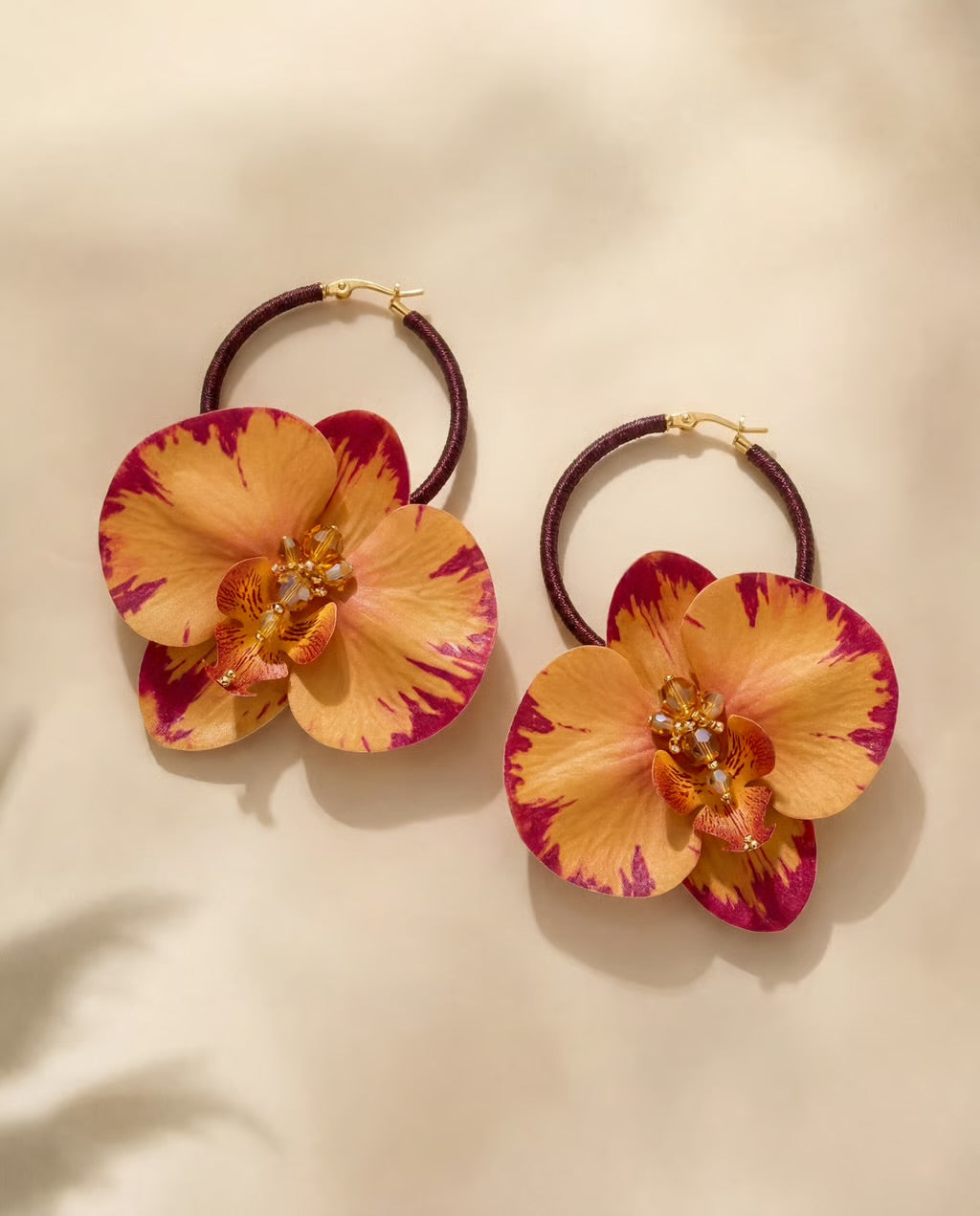 A pair of hoop earrings featuring large marbled orange and magenta orchid flowers with gold-tone hardware, displayed on a warm beige background.