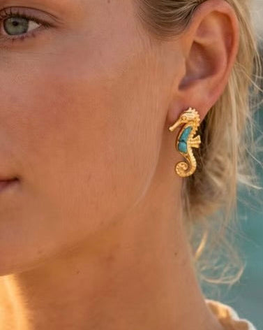 A close-up of a woman's ear wearing a small gold seahorse-shaped stud earring with a turquoise blue inlay.