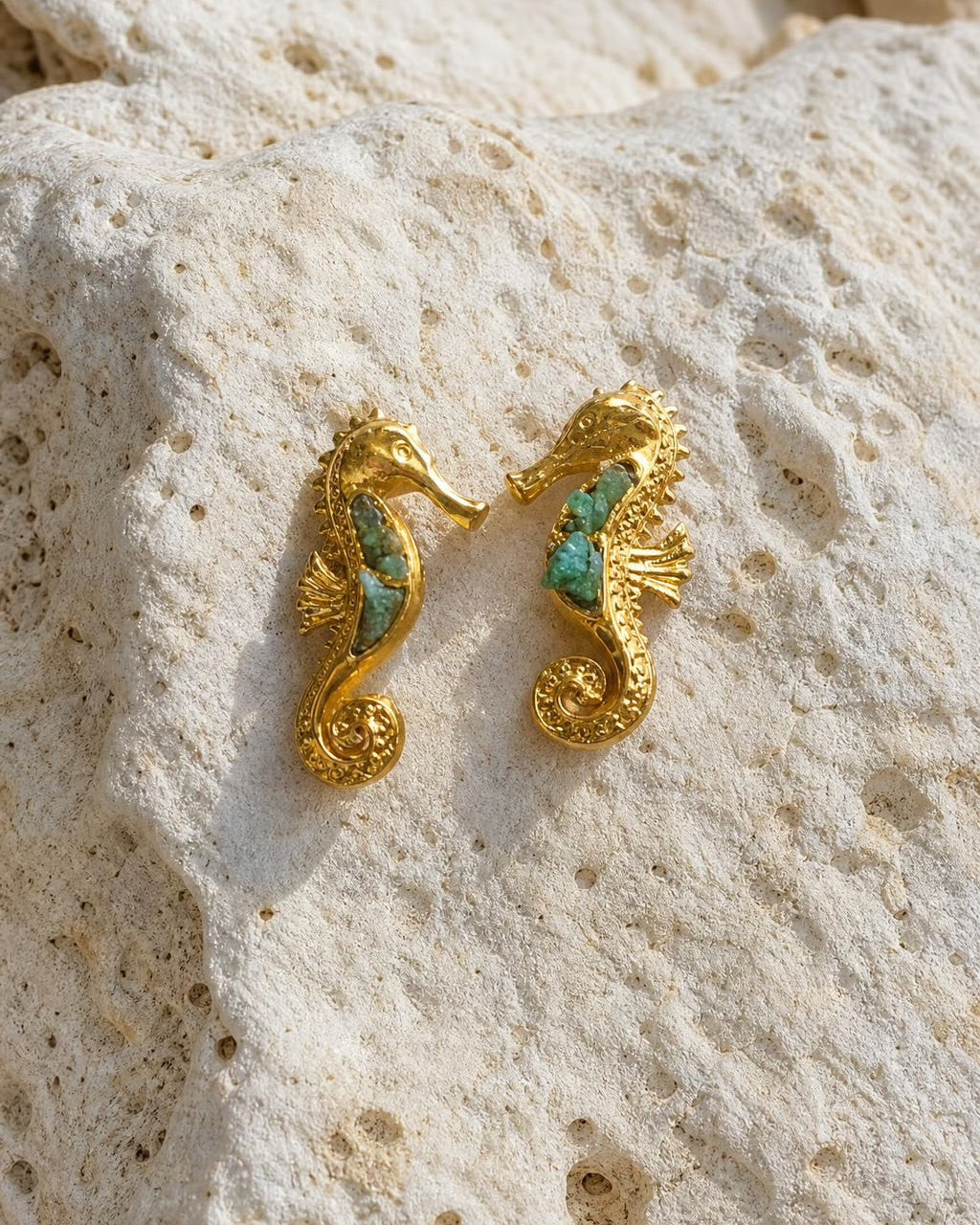 A pair of gold seahorse-shaped earrings with turquoise inlay, resting on a textured beige stone surface.