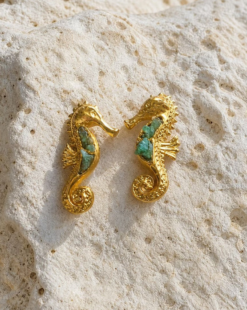 A pair of gold seahorse-shaped earrings with turquoise inlay, resting on a textured beige stone surface.