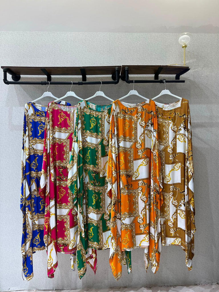 Four long printed kaftan-style dresses or sarongs hang on white hangers from a black wall rack against a light gray wall. The garments feature ornate baroque-style motifs in different colorways: blue and white, pink and white, green and white, and orange and white, each with gold detailing.