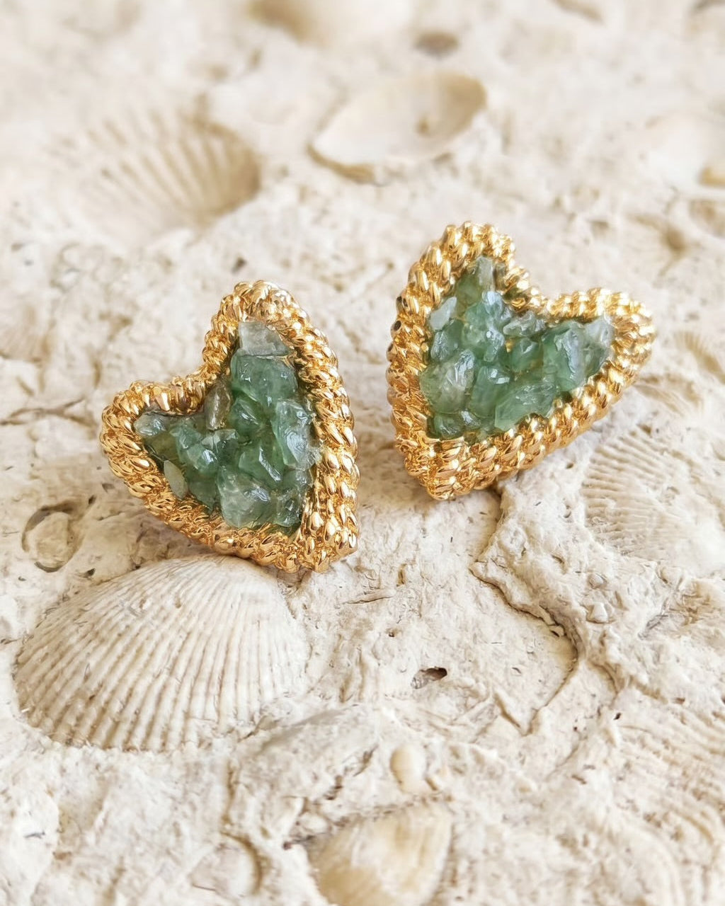A pair of heart-shaped gold stud earrings with green crystal inlays, displayed on a textured beige stone surface with seashell motifs.