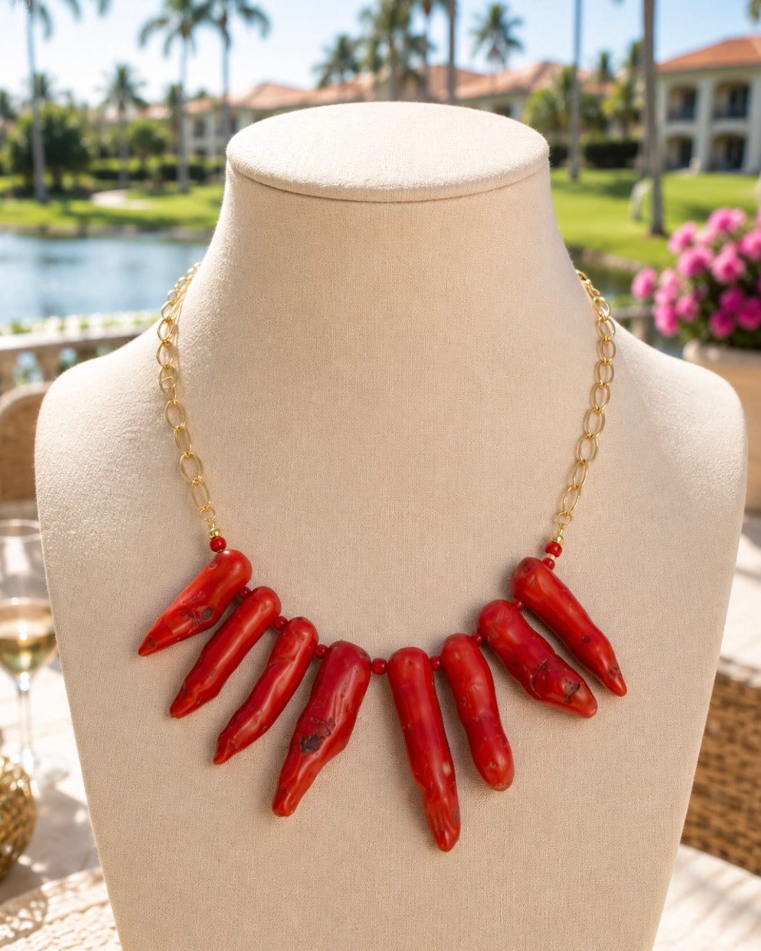 A statement collar necklace made of ten red conical beads attached to a gold chain, displayed on a cream-colored mannequin bust with an outdoor resort background.