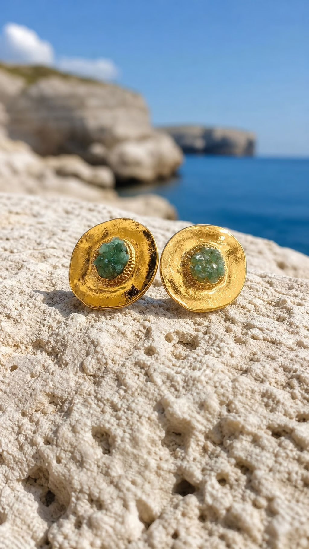 A pair of gold round stud earrings with green stone centers, resting on a textured white rock with a blue sea and rocky coastline in the background.