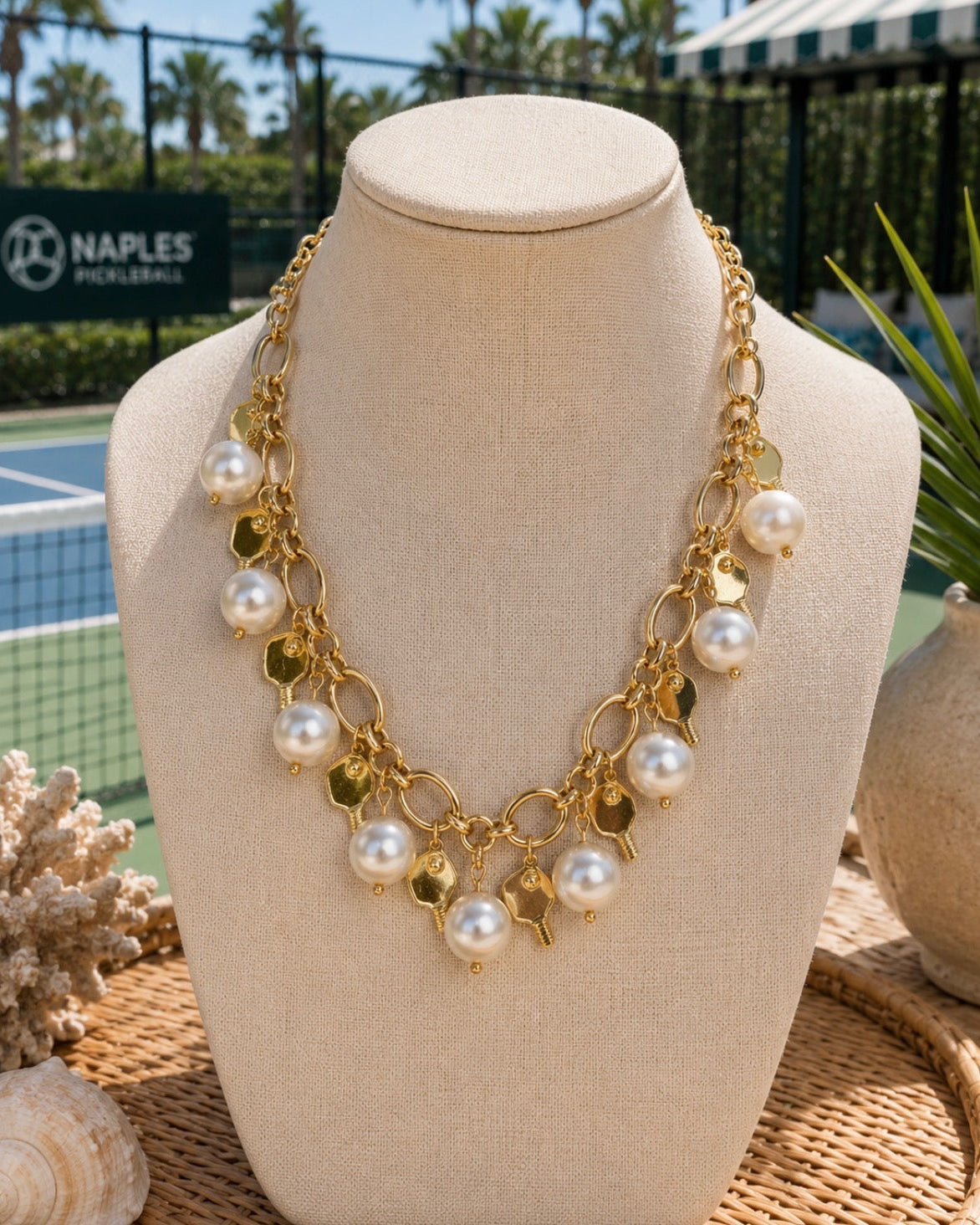 A gold statement necklace with faux pearls and gold leaf charms displayed on a beige fabric mannequin bust, set against a blurred tennis court background with a Naples Pickleball sign.