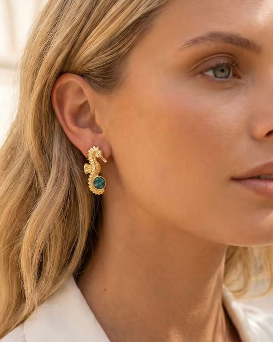 A close-up of a woman wearing a gold seahorse-shaped dangle earring with a green oval stone, set against a soft, light background.