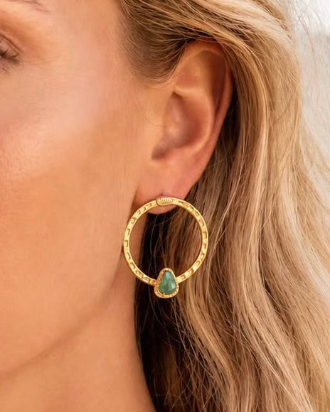 A close-up of a woman wearing a textured gold hoop earring with a small green teardrop gemstone dangling from the side.