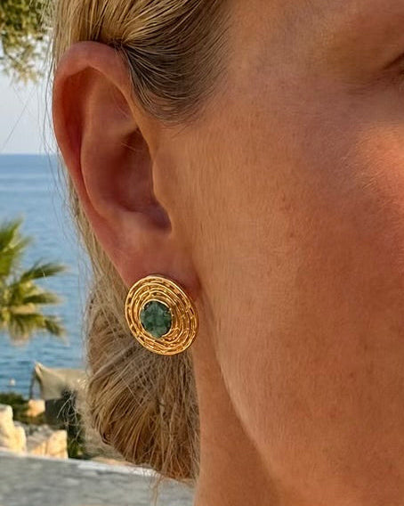 A close-up of a woman's ear wearing a gold circular stud earring with a green circular center, set against a blurred outdoor background of water and trees.