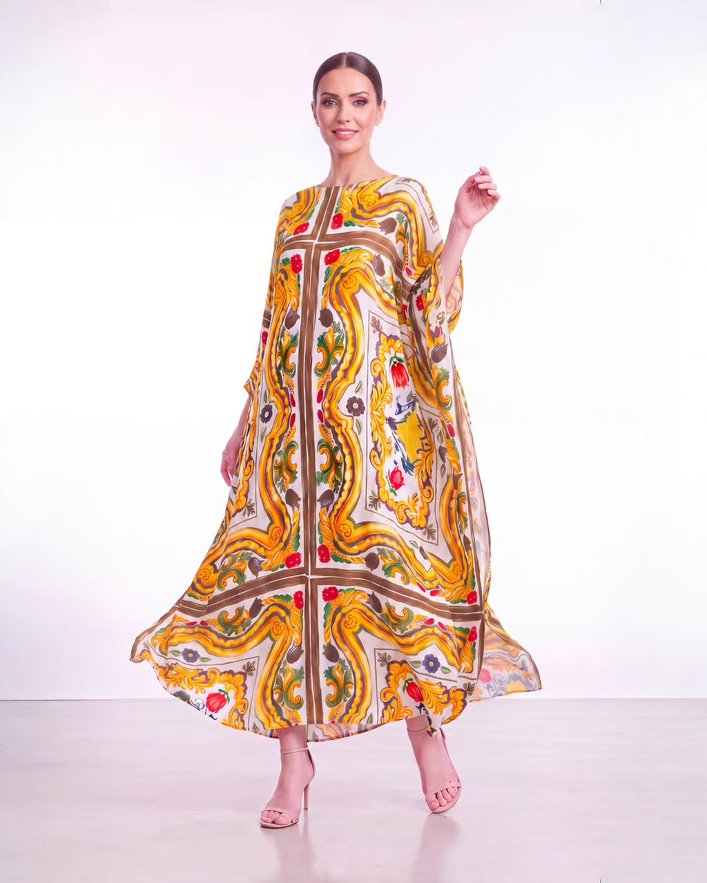 A woman modeling a flowing kaftan dress with a vibrant yellow, orange, and brown paisley pattern on a white background.